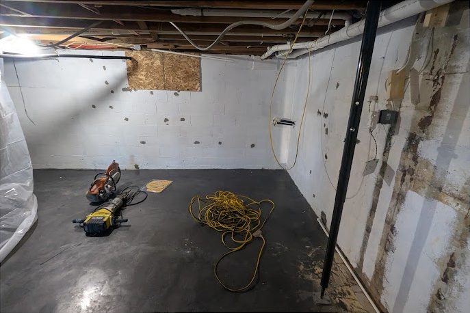 Before: a basement corner mid-job, with a concrete saw, a demolition hammer and a coiled cord on the floor