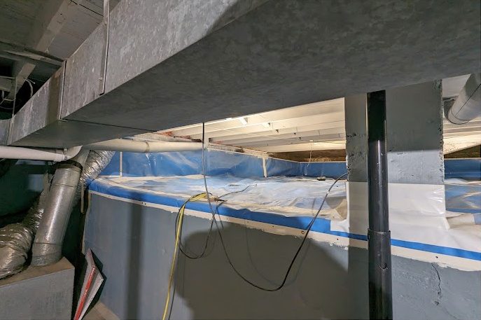 A crawl space behind a basement wall, lined, with blue tape along the liner edge
