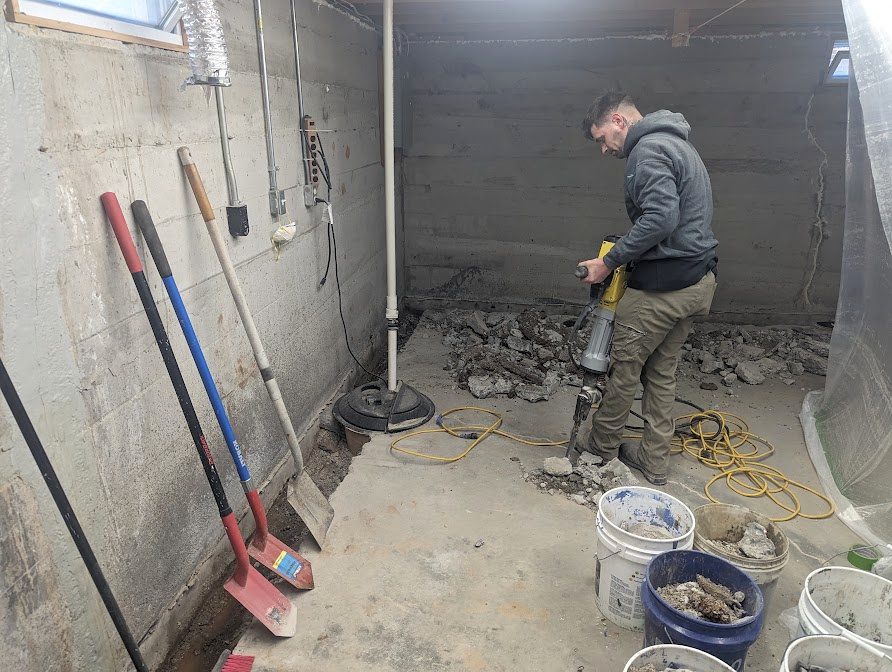 A crew member opening the floor along a basement wall, beside the sump area.