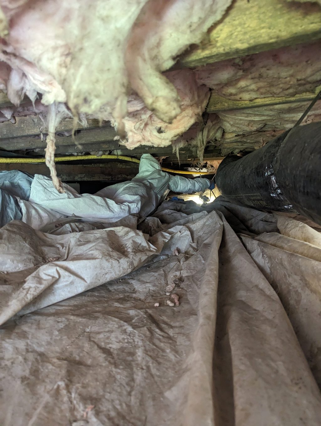 Low crawl space with loose material on the ground and insulation hanging from the joists