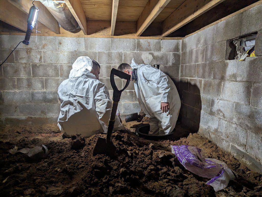 Two coveralled crew members fitting a basin in crawl space corner